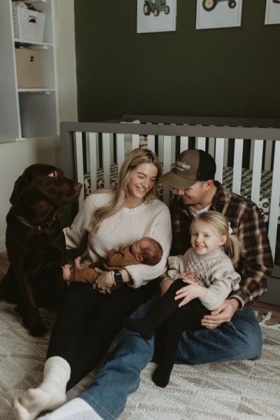 Samantha and her husband sitting on a nursery floor with their newborn baby, toddler daughter, and family dog.